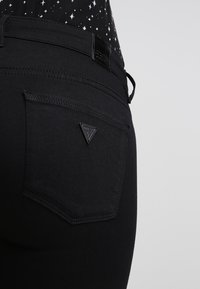 Černé džíny z denimu s texturovaným povrchem, které mají trojúhelníkové logo "Guess" na zadní kapse a hladký pas.