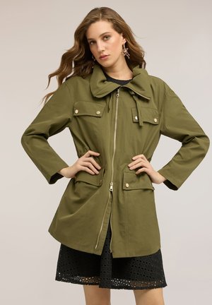 Donna con capelli mossi che indossa una giacca verde oliva con zip, quattro tasche a patta e bottoni dorati sopra un vestito nero di pizzo, con le mani sui fianchi.