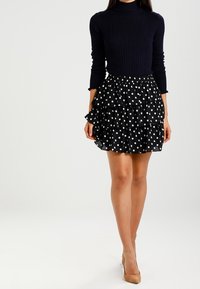 Nainen, jolla on päällään musta ribattua neuletta oleva poolopaita ja musta kerroksellinen hame, jossa on valkoisia pilkkuja, yhdistettynä beigeihin korkokenkiin.