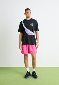 Riñonera en lavanda y menta, fabricada en material ligero, llevada cruzada sobre el torso. Camiseta negra, pantalones cortos rosas y zapatos negro-azul.