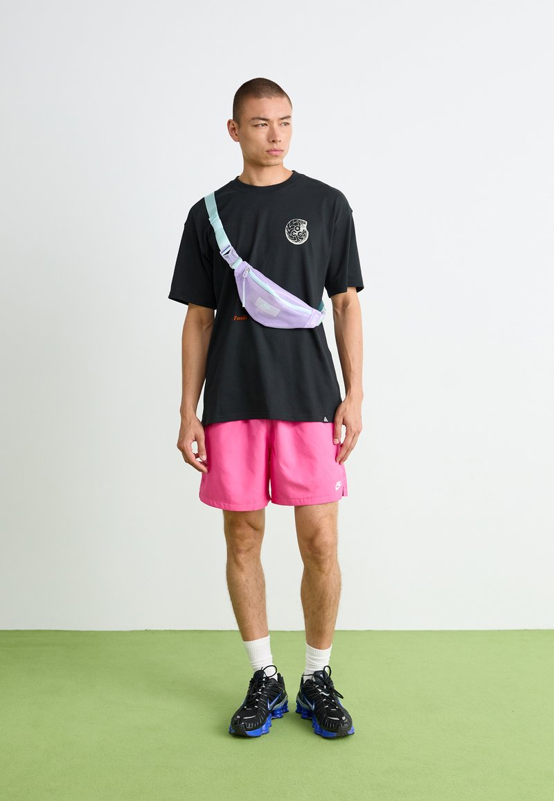 Sac banane en lavande et menthe, fait de matériau léger, porté en travers du torse. T-shirt noir, shorts roses et chaussures noir-bleu.