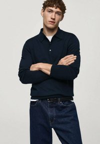Polo a maniche lunghe blu navy con colletto, abbinato a jeans in denim scuro con vestibilità slim e texture sottile, indossato con una cintura nera.