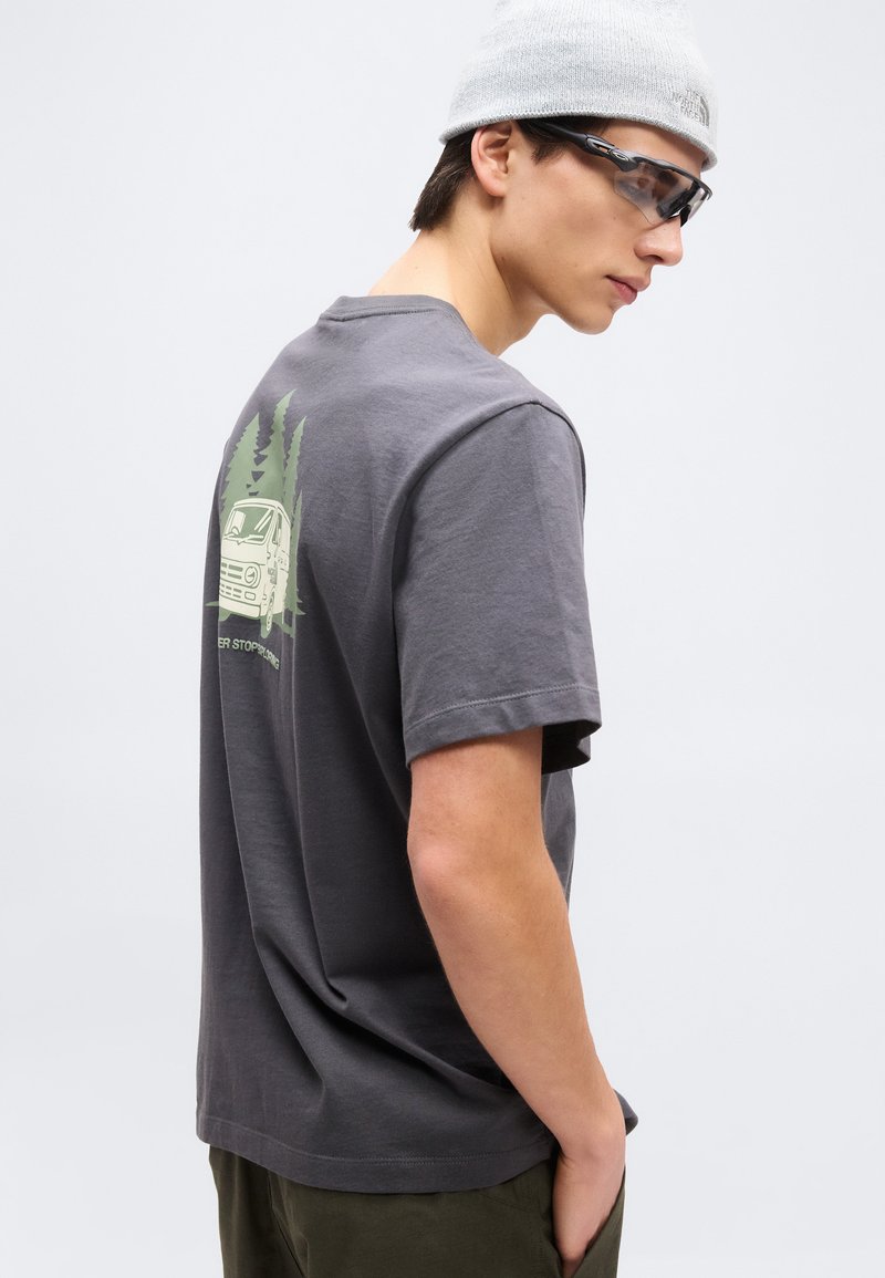 Junger Mann, der ein graues T-Shirt mit einem Grafikaufdruck von einem Van und einem Baum auf dem Rücken, eine graue Mütze, dunkle Sonnenbrille und grüne Hose trägt, schaut seitlich.