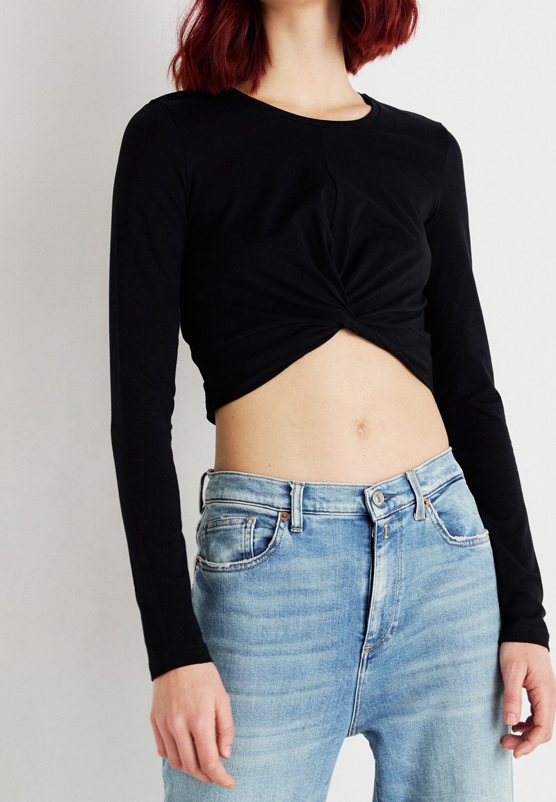 Zwarte langmouw cropped top met een geknotte voorkant, gecombineerd met lichtblauwe high-waisted jeans met een vervaagt textuur.