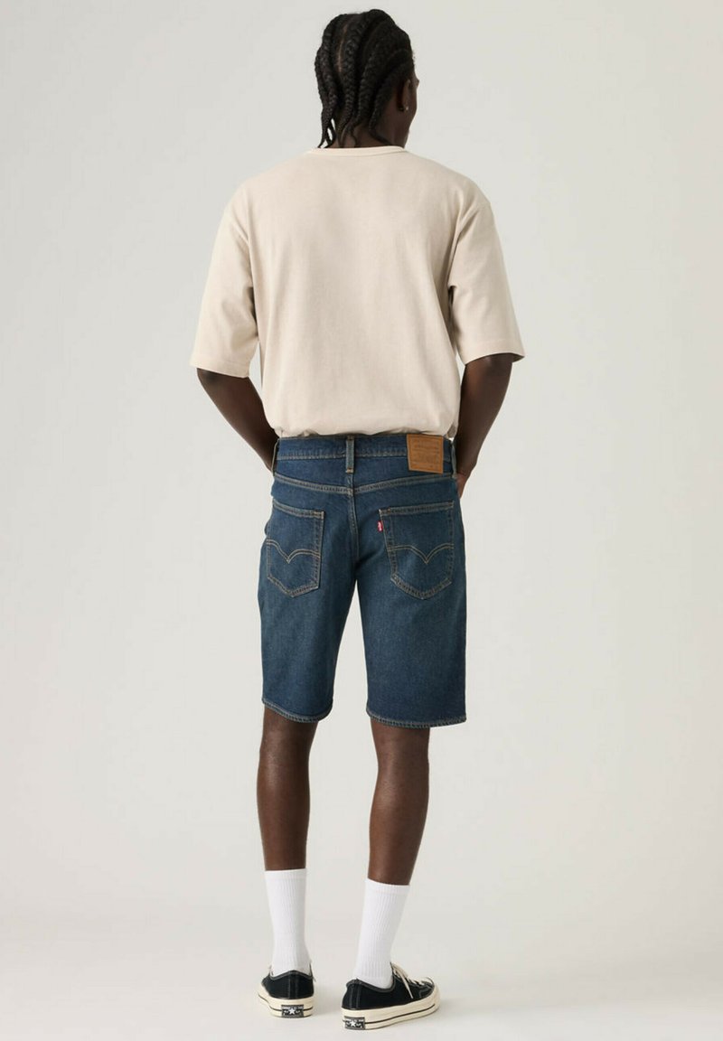 Homme aux cheveux tressés portant un t-shirt beige surdimensionné, un short en denim bleu, des chaussettes blanches et des baskets basses noires, debout de dos.