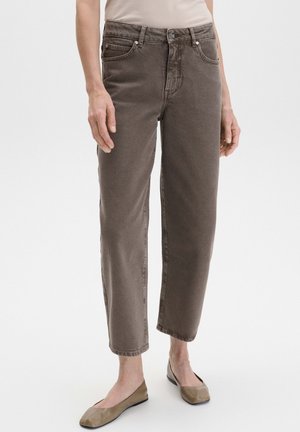 Femme portant un jean taupe droit et des chaussures plates beiges, debout sur un fond blanc uni.