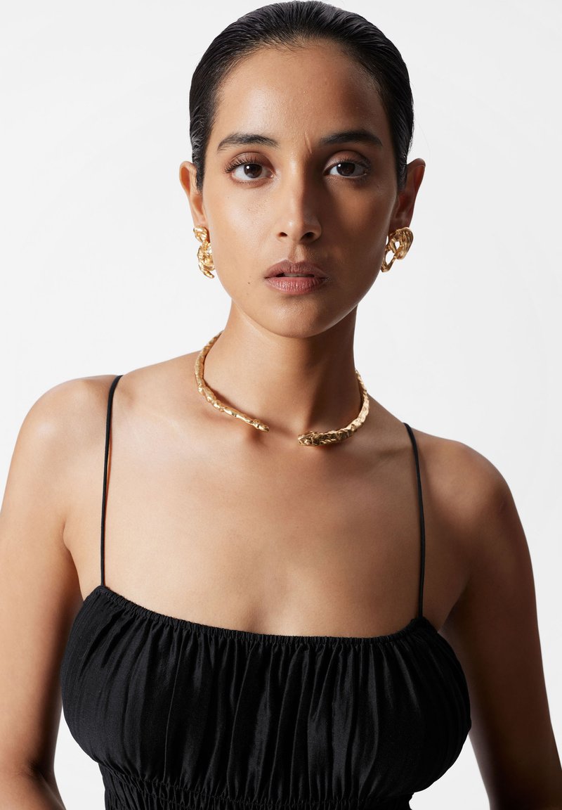 Mujer con cabello oscuro y liso, que lleva pendientes de oro texturizados y un collar de brazalete abierto, además de una blusa negra de tirantes, frente a un fondo blanco liso.