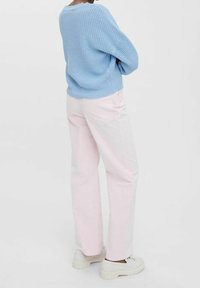 Osoba w jasnoniebieskim swetrze z dzianiny, jasnoróżowych, szerokich spodniach i białych butach na platformie, stoi z założonymi na piersi rękoma na białym tle.