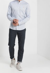 Mies, joka säätää vaaleansiniseksi napitettuja paitoja, pukeutuneena tummiin kuvioituihin housuihin, jotka on kääritty nilkkojen ympärille, ja vaaleisiin tennareihin, seisoo valkoista seinää vasten.