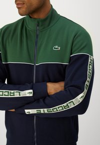 Homem a usar uma jaqueta com fecho éclair verde e azul-marinho, com riscas brancas e verdes no comprimento das mangas e um pequeno logótipo de crocodilo verde no peito.