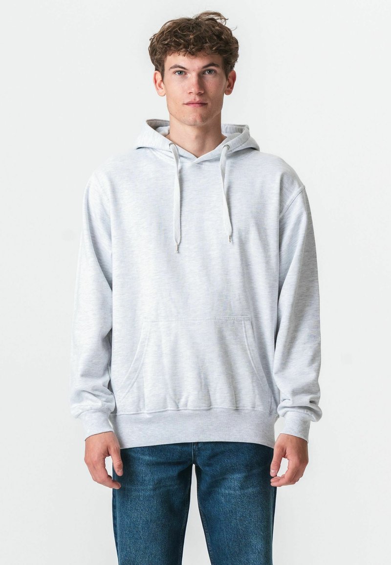 Jeune homme aux cheveux bouclés portant un sweat à capuche gris clair et un jean bleu, debout devant un fond blanc.