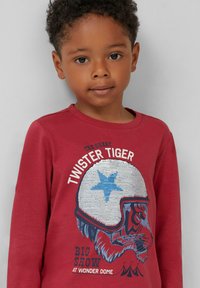 Rotes Langarmshirt mit einem Tiger-Design, der einen weißen Helm mit einem blauen Stern trägt. Der Text lautet: "TWISTER TIGER BIG SHOW IM WONDER DOME."