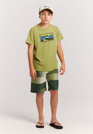 Groen katoenen T-shirt met golfgrafiek, gecombineerd met groen gestreepte zwembroek. Zwarte slippers en een witte pet met blauw logo maken de outfit compleet.