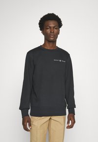 Felpa nera in cotone con scollo rotondo, polsini e orlo a costine, caratterizzata dal logo "GANT 1949" sul petto. Indossata sopra pantaloni color khaki.