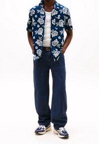 Short-sleeve navy floral shirt, white tank top underneath, dark blue baggy pants, and navy sneakers with white accents.