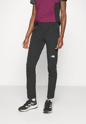 Person trägt schwarze The North Face Hose, schwarz-weiße Sneaker und ein kurzärmeliges bordeaux-schwarzes Shirt und steht vor einem weißen Hintergrund.