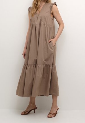 Vrouw draagt een mouwloze midi-jurk in taupe met geruite schouderdetails en een gelaagde rok, staand met één hand in een zak.