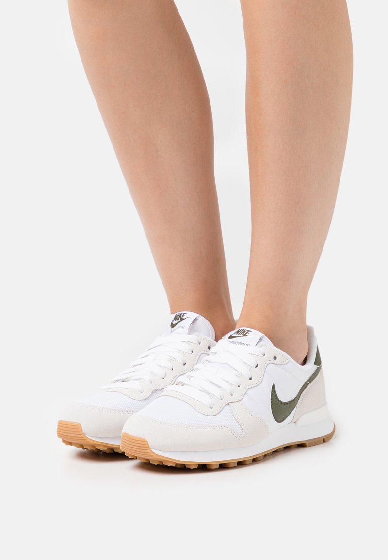 Paire de baskets blanches Nike avec un swoosh vert, portées pieds nus, debout sur une surface blanche.