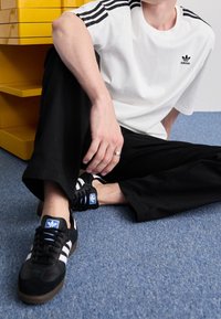 Bílý tričko s černým designem tří pruhů, černé kalhoty s širokými nohavicemi a černé tenisky s bílými pruhy a modrými akcenty.