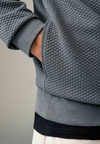 Grijze sweatshirt met textuur en een ruitpatroon, geribbelde mouwen en een tailleband. Zichtbare zak aan de zijkant, gelaagd over een marineblauw shirt.