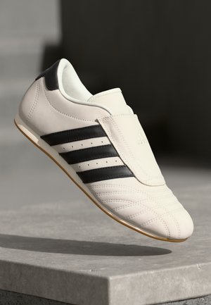 Baskets de sport en cuir blanc avec des rayures noires, bout arrondi, col rembourré et détails texturés, sur un fond neutre.