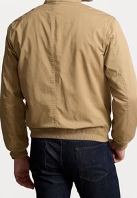 Beige bomberjack met een gladde textuur, geribbelde manchetten en zoom, en een eenvoudig ontwerp. Gedragen over donkere spijkerbroek, gezien van achteren.