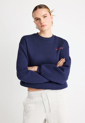 Granatowa bluza dresowa z okrągłym dekoltem, luźnym krojem i ściągaczami na rękawach. Na piersi widoczne dyskretne czerwone logo. Noszona z szarymi spodniami dresowymi.