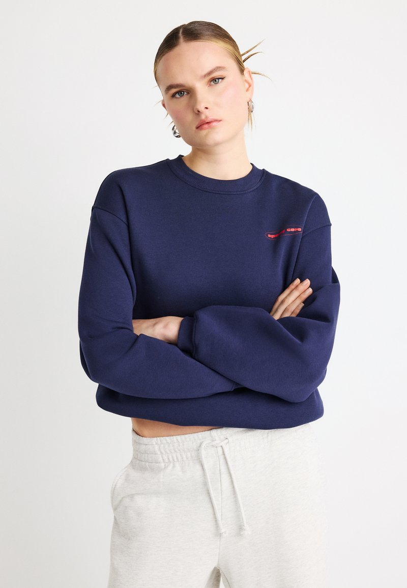 Tumesinise värvusega pusa, millel on ümara kaelusega, lõdva lõike ja soonikkoes varrukasuu. Esiosal on õrn punane logo. Kantud hallide dressipükstega.