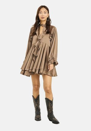 Mujer con un vestido suelto marrón con volantes y mangas largas combinado con botas de vaquero negras bordadas sobre un fondo liso.