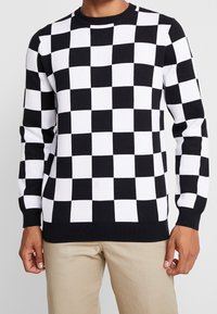 Homme portant un pull à manches longues noir et blanc à carreaux avec un pantalon beige, se tenant devant un fond clair.