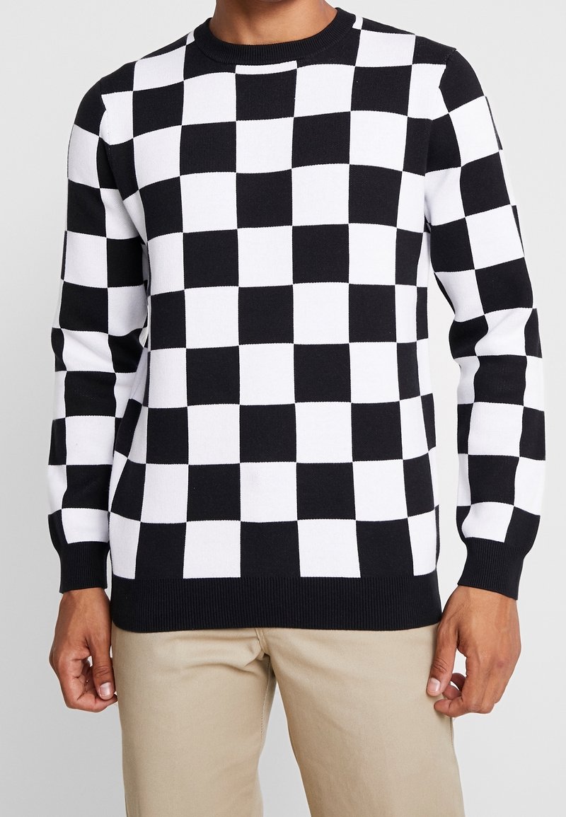 Homme portant un pull à manches longues noir et blanc à carreaux avec un pantalon beige, se tenant devant un fond clair.