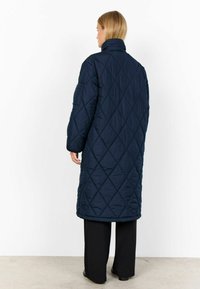 Quiltad marinblå lång kappa med hög krage, med diamantformad sömnad och lös passform. Svarta byxor och skor synliga nedanför.