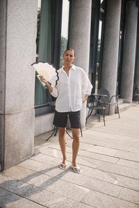 Chemise oversized blanche perforée, shorts noirs et tongs blanches. Tenant un éventail décoratif avec un bord festonné. Pavage en pierre en arrière-plan.