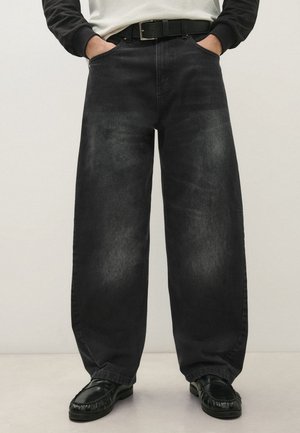 Personne portant un jean large noir délavé, ceinture noire, mocassins noirs et une chemise blanche et noire à manches longues, les mains dans les poches.