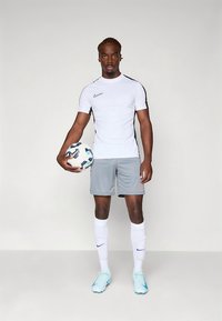 Camisa branca de mangas curtas com detalhes pretos, calções cinzentos, meias brancas e chuteiras azul claro; segurando uma bola de futebol com padrão.