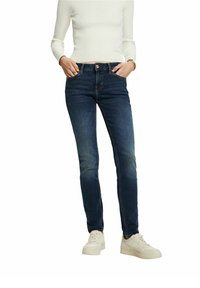 Personne portant un jean slim bleu foncé, un haut côtelé à manches longues blanc, des baskets blanches, les mains dans les poches avant sur fond blanc.