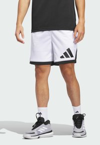 Weiße Sportshorts mit schwarzem Rand und drei schwarzen Streifen auf der linken Seite, kombiniert mit grauen Turnschuhen und weißen Kniestrümpfen.