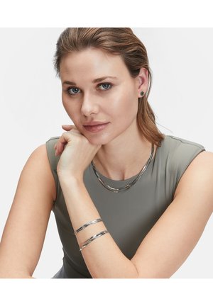 I gioielli in argento presentano tre pezzi metallici lisci e lucidi, tra cui una collana e due bracciali, indossati da un modello con pelle chiara.
