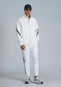 Weißer Zip-Jacket mit schwarzen Seitestreifen, dazu passende weiße Hose und graue Sneakers. Besitzt einen hohen Kragen und eine lockere Passform.