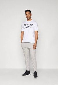 Vyras, vilkintis baltą marškinėlį su Reebok logotipu, šviesiai pilkas sportines kelnes ir juodus sportinius batus, stovi priešais šviesų vienodą foną.