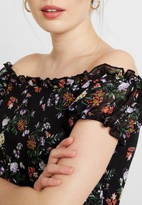 Svart blomsteroff-shoulder topp med volangärmar, med en blandning av livfulla blommor i lila, rött och vitt mot en mörk bakgrund.