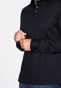 Svart långärmad polo med krage och knappar, med en mjuk struktur och nätta ärmslut. Tyget verkar mjukt och lätt.