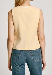 Top senza maniche in morbido tessuto giallo con scollatura quadrata, caratterizzato da una cucitura discreta sul retro e un'etichetta sotto il collo. Abbinato a jeans blu.