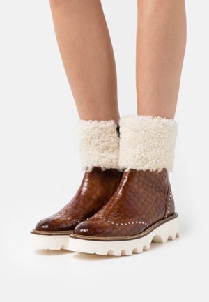 Botas de tobillo de cuero marrón con textura de cocodrilo, puño de borreguito blanco, suela de plataforma blanca gruesa y acentos de tachuelas decorativas.