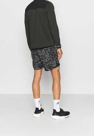 Mann trägt ein schwarzes und dunkelgrünes Langarmoberteil, graue Shorts mit Leopardenmuster, weiße Adidas-Socken und schwarze Turnschuhe, steht mit dem Rücken zur Kamera.