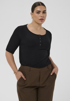 Vrouw met donker haar in een staart, gekleed in een zwart topje met knopen en ellebooglange mouwen en een bruine broek, handen in de zakken, neutrale achtergrond.