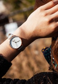 Montre ronde avec un cadran argenté, des accents en or rose et un bracelet en cuir gris clair, présentant des index et des aiguilles minimalistes.