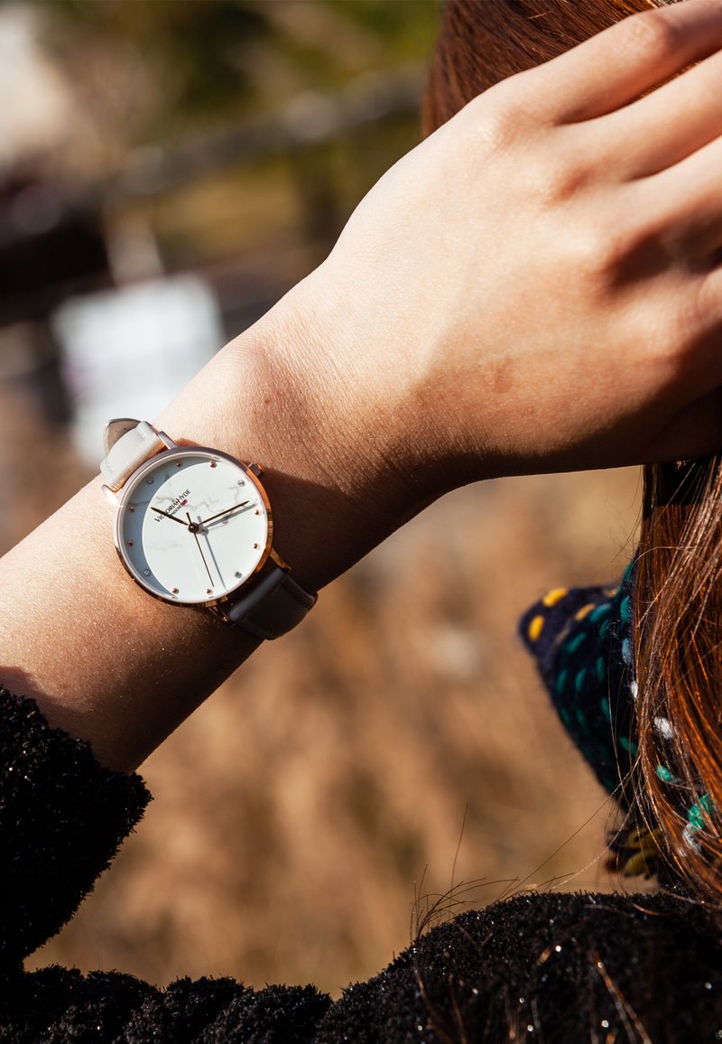 Montre ronde avec un cadran argenté, des accents en or rose et un bracelet en cuir gris clair, présentant des index et des aiguilles minimalistes.