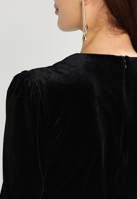 Vestido de terciopelo negro con escote redondeado y mangas abullonadas. Presenta una cremallera en la parte posterior y una textura suave y lisa. Gran pendiente dorado visible.