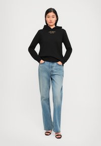 Hoodie noir avec un logo sur le devant, associé à un jean droit bleu clair. Le modèle porte des sandales noires et se tient les mains dans les poches.